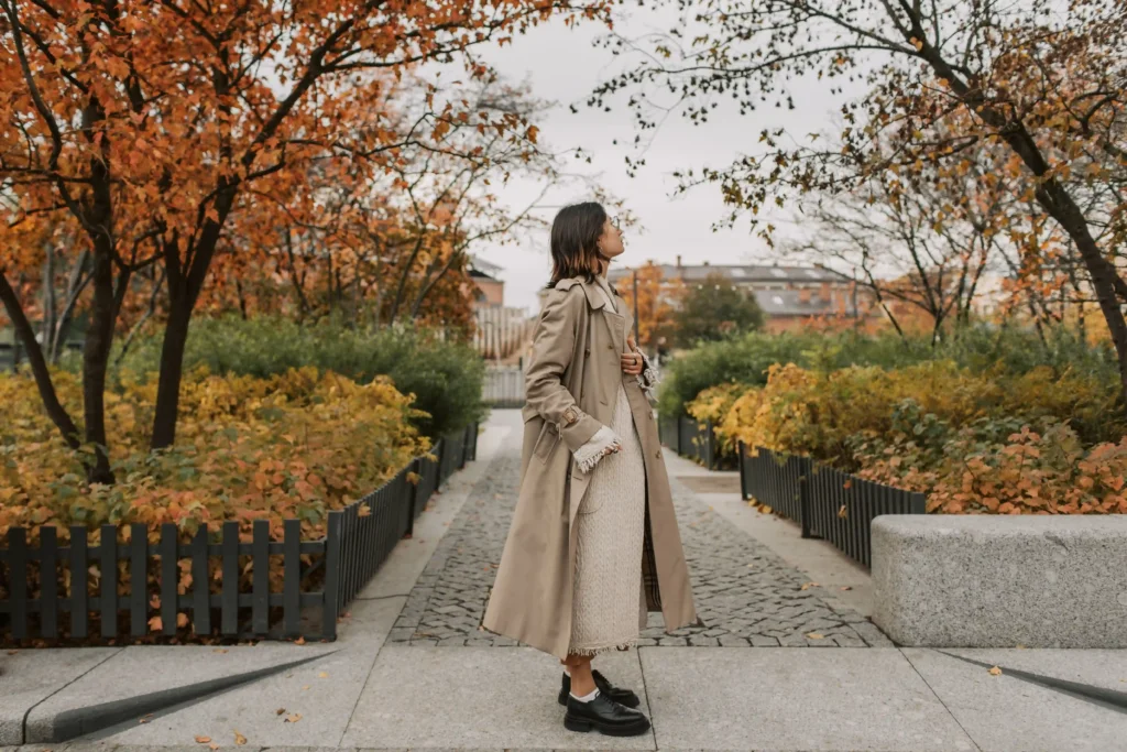 Woman wearing a beige trench coat over a cream knit midi dress with black loafers