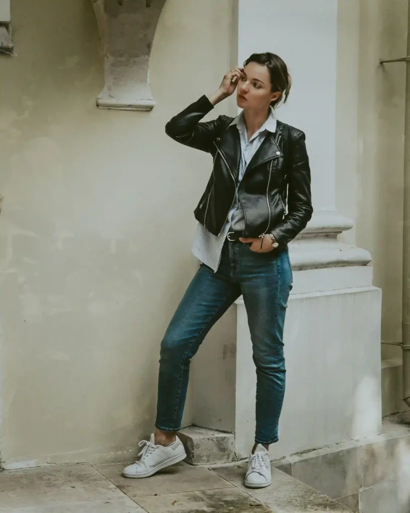 Woman wearing a black biker-style leather jacket, blue jeans and white sneakers