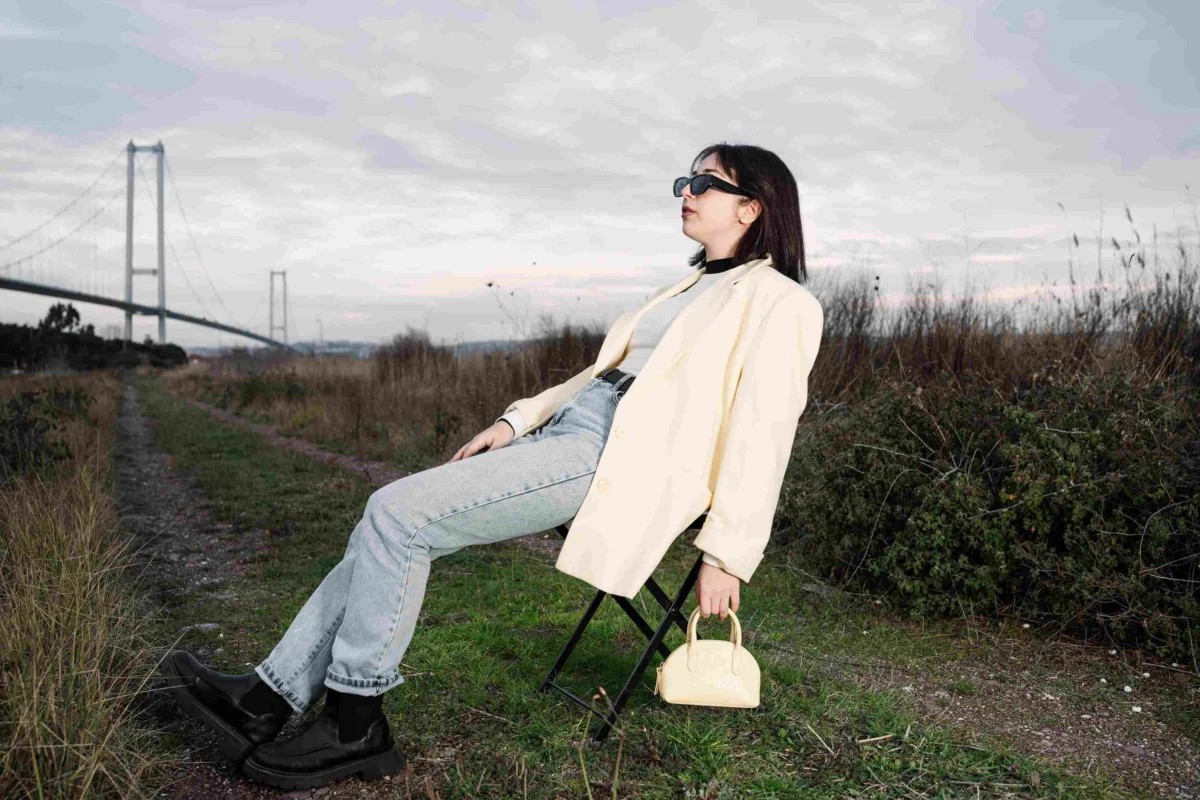Woman wearing a cream blazer, light blue jeans, black ankle boots and sunglasses while sitting outdoors.