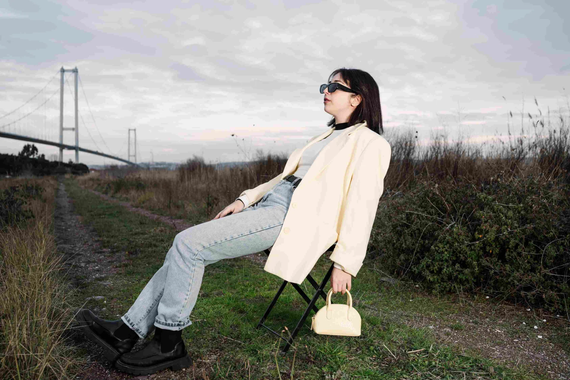 Woman wearing a cream blazer, light blue jeans, black ankle boots and sunglasses while sitting outdoors.