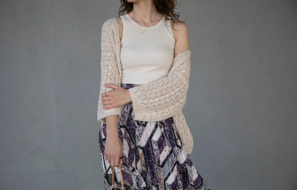 Woman wearing a cream open-knit cardigan over a white tank top with a purple patterned skirt