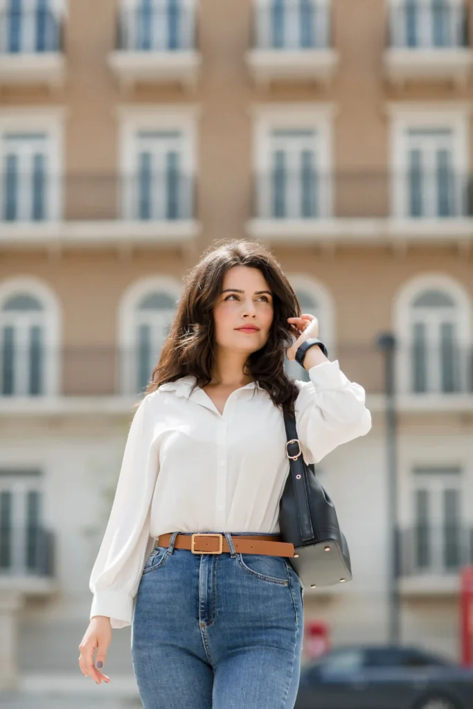Woman wearing a white blouse, blue jeans, tan belt and black shoulder bag on a city street.