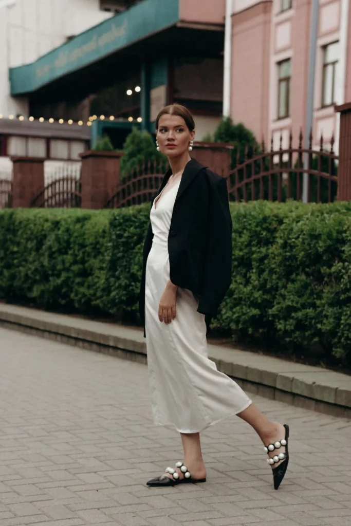 Woman wearing a white midi dress with a black jacket over her shoulders and pointed flats